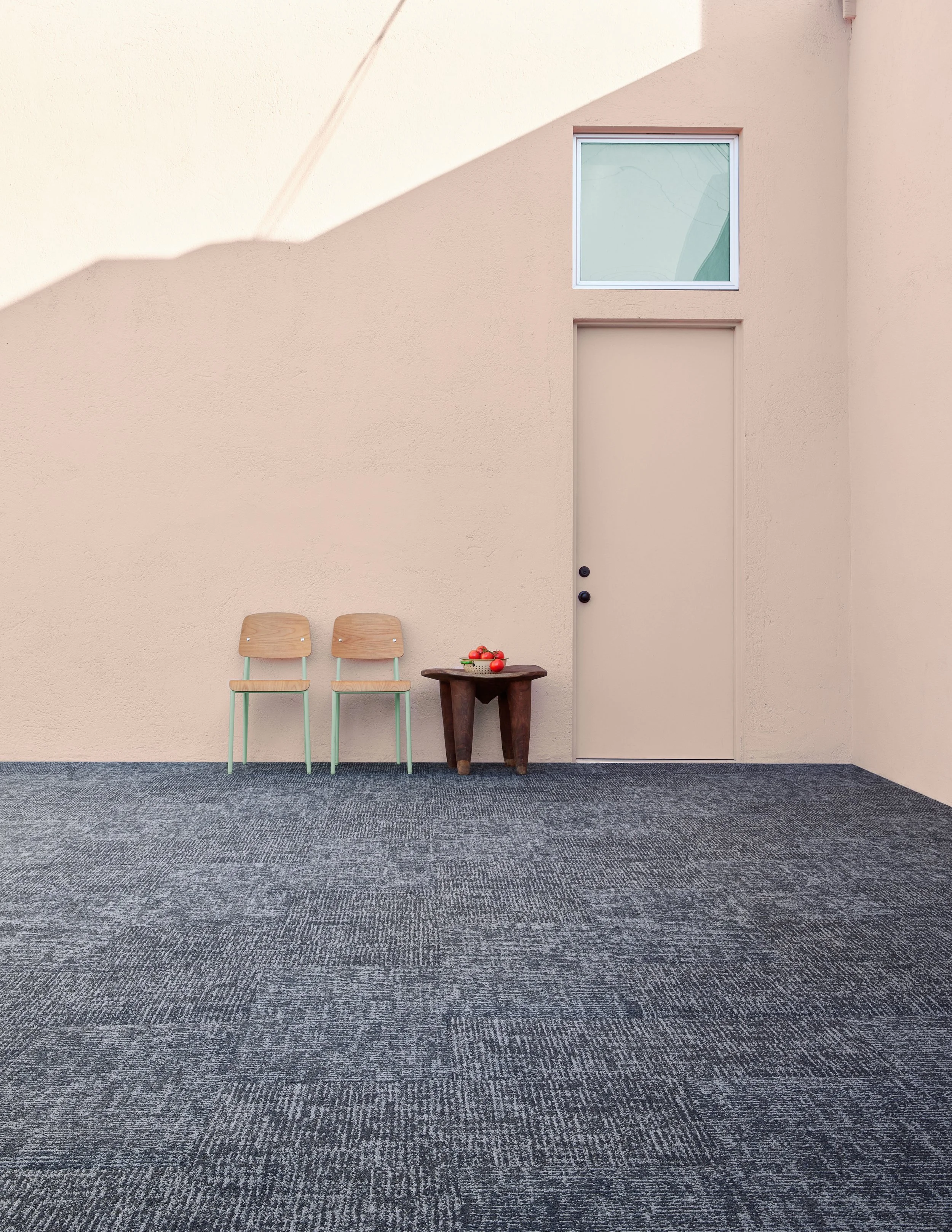 Two wood chairs and a dark stool with tomatoes against a peach wall on textured gray flooring.