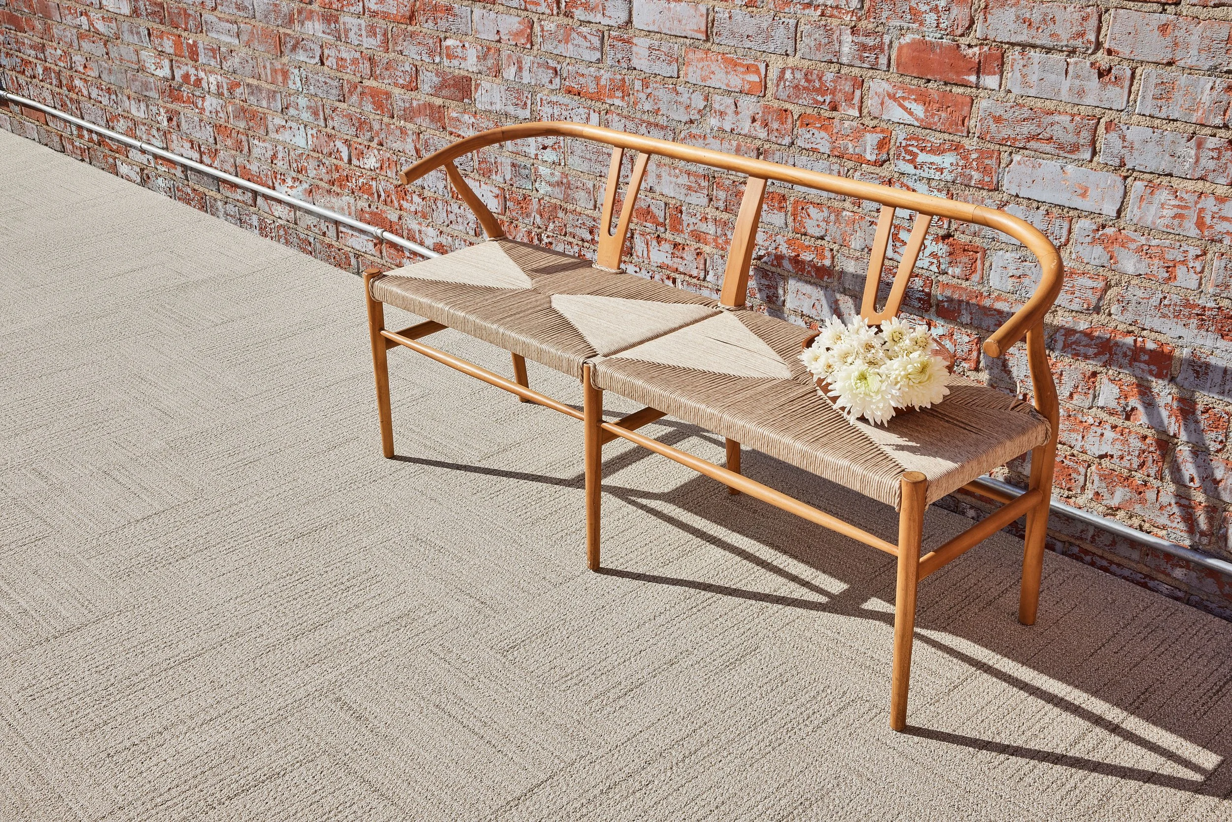 Light wood bench with a woven seat and white flowers against a brick wall in direct sunlight.