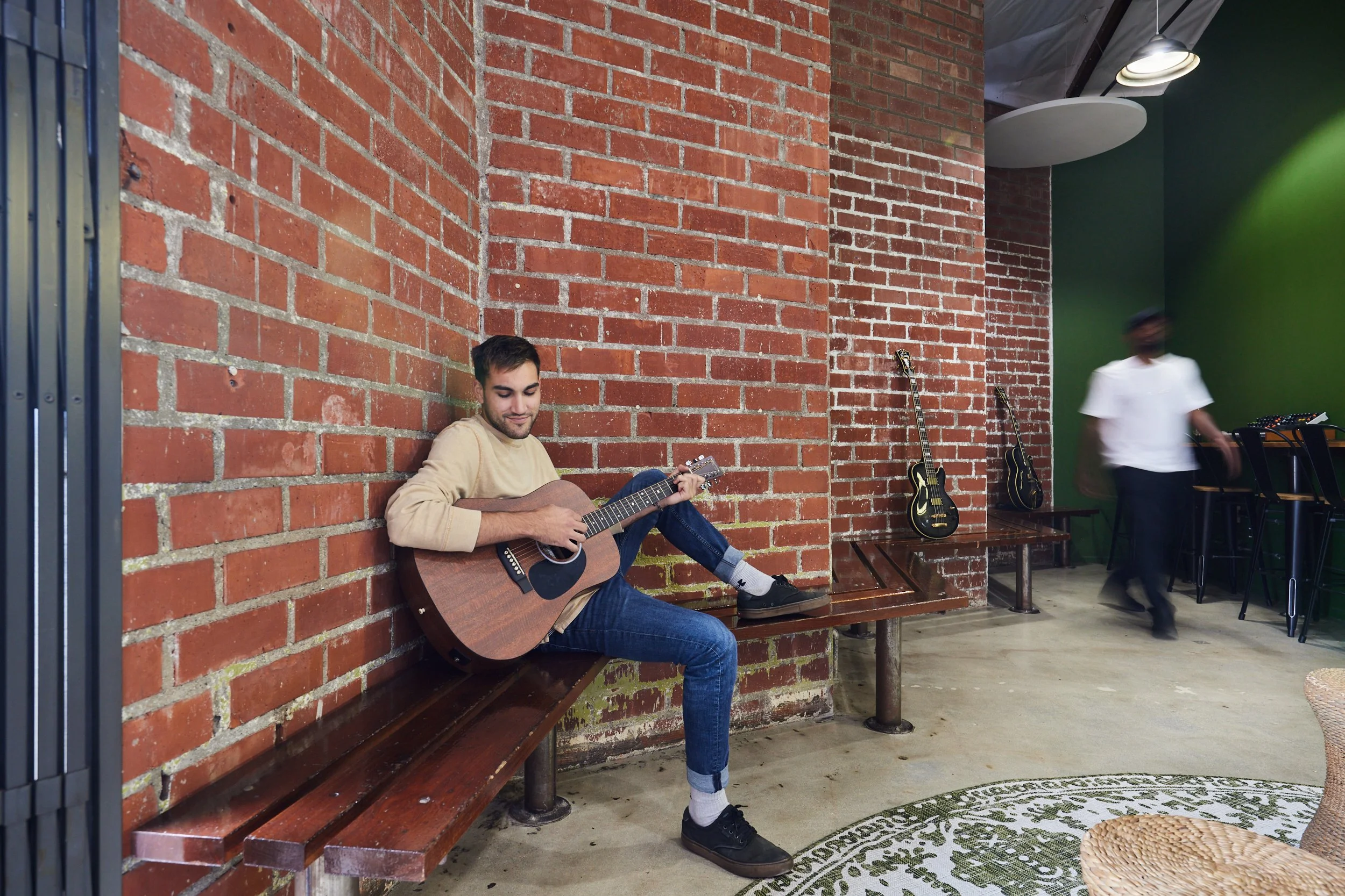 Man playing an acoustic guitar on a wood bench against a red brick wall in bright indoor light.