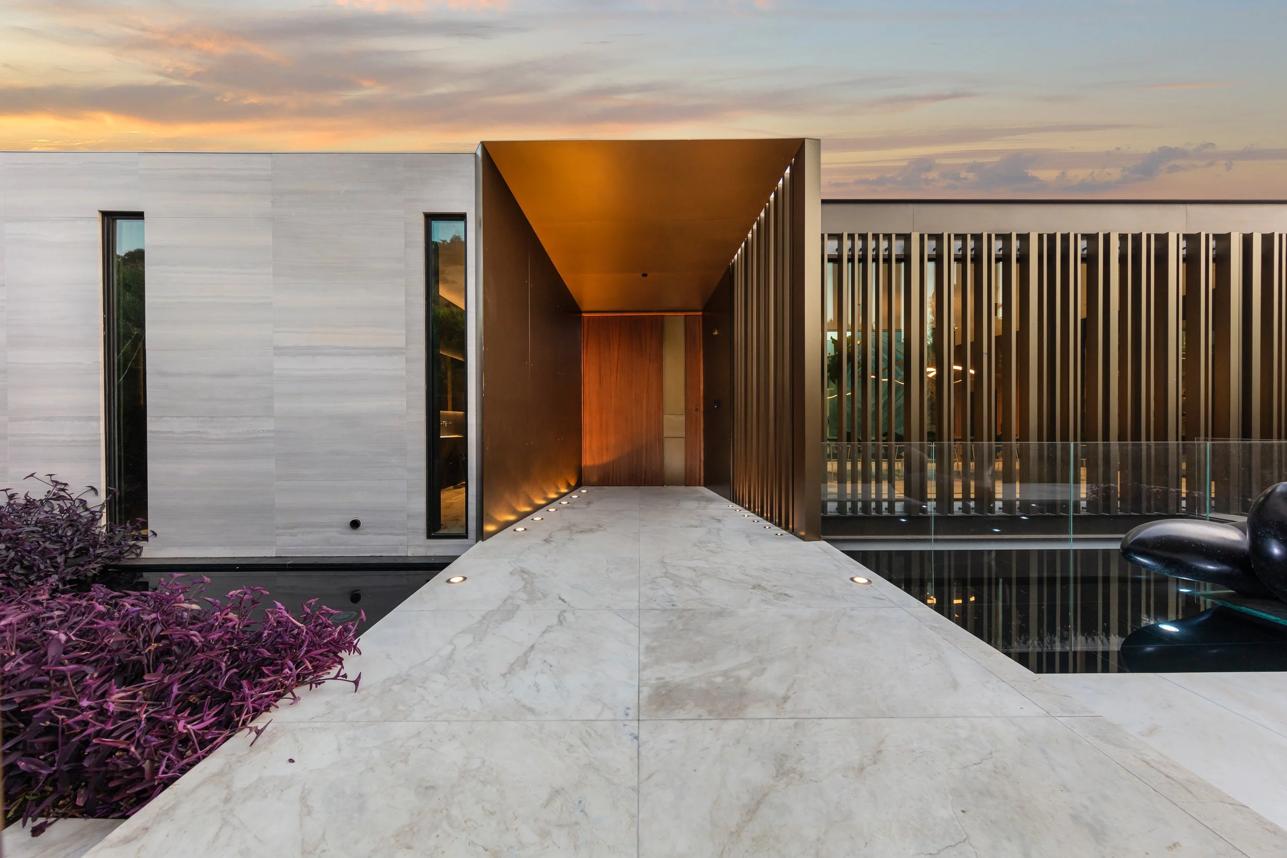Lit bronze entry canopy, marble walkway, and reflecting pool at a modern residence at dusk.