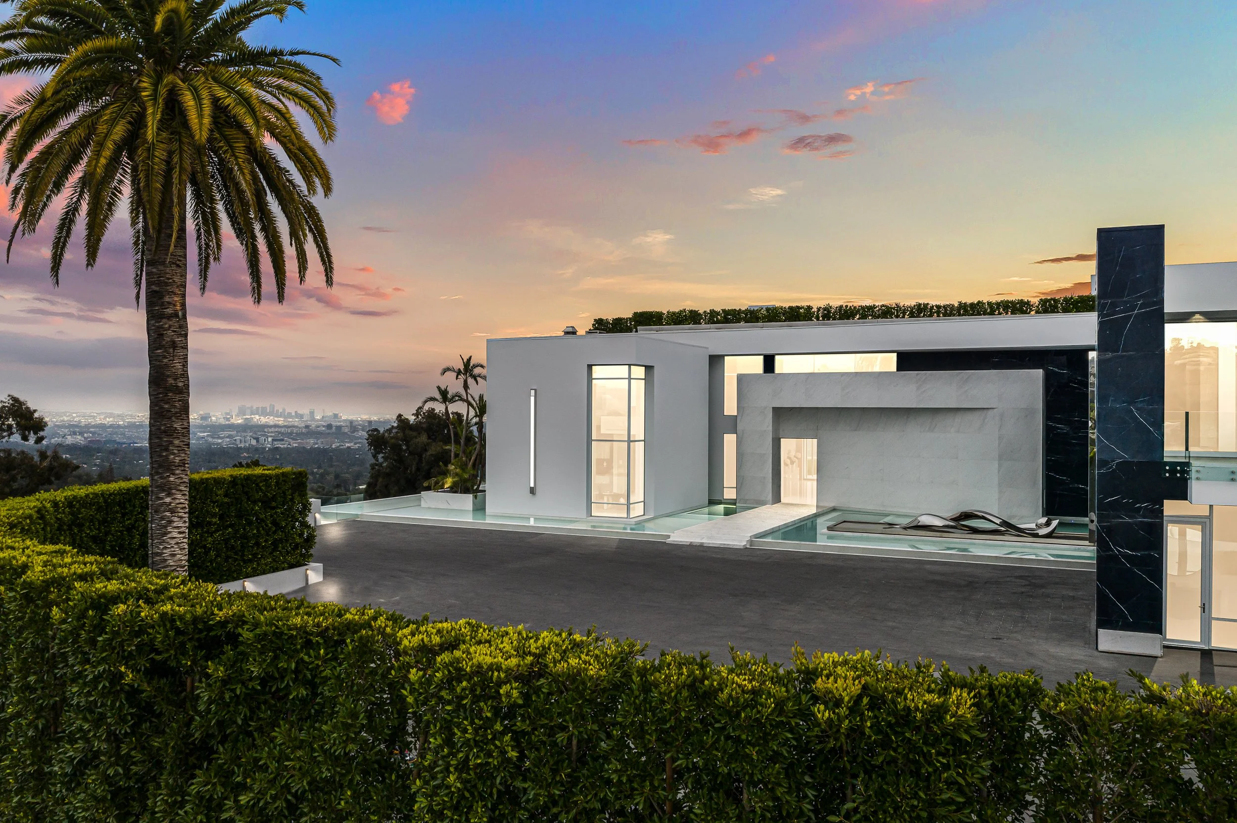 White hillside residence at dusk with reflecting pools, a palm tree, and a distant city skyline.