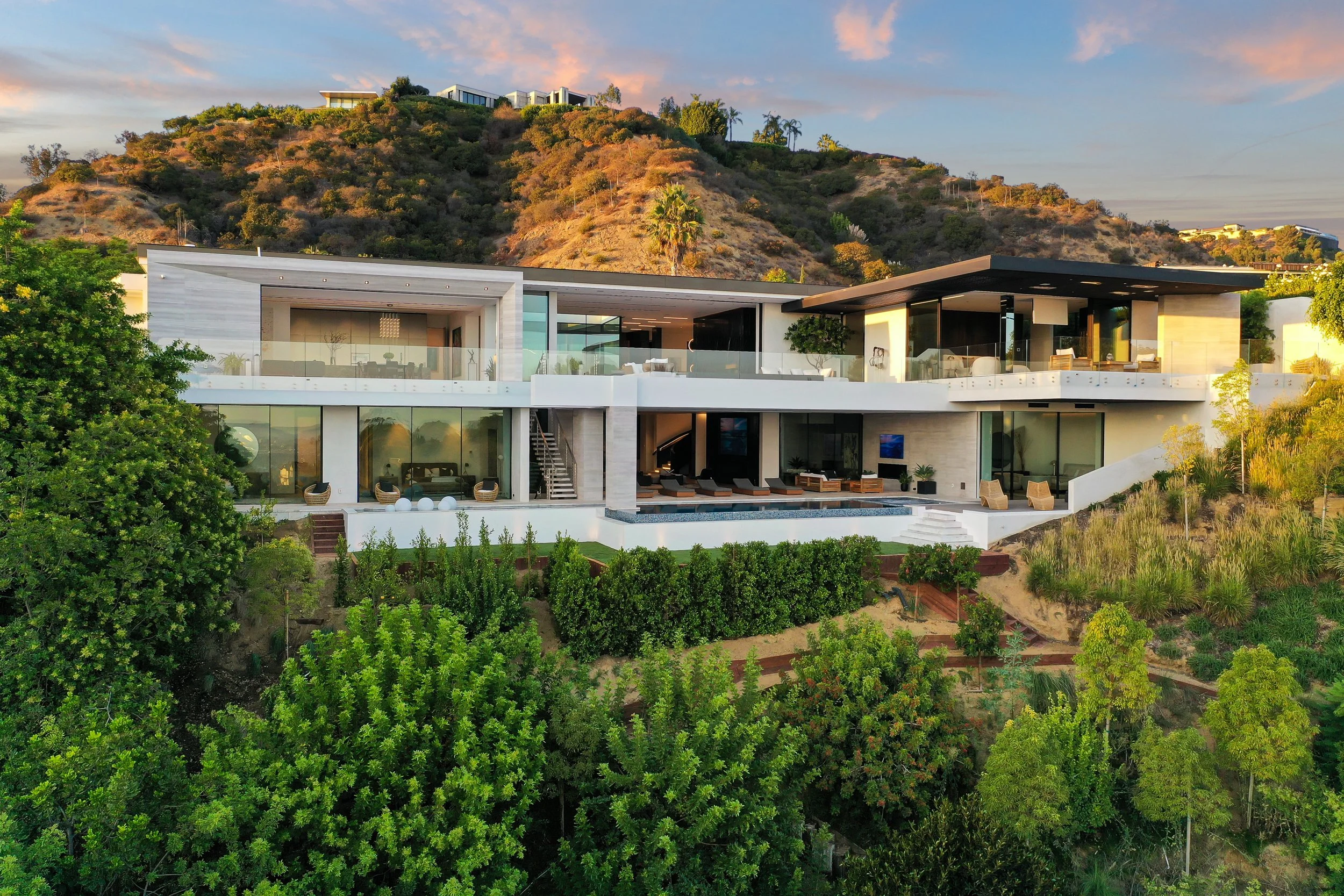 White multi-story hillside residence with glass walls and a terrace pool under warm sunset light.