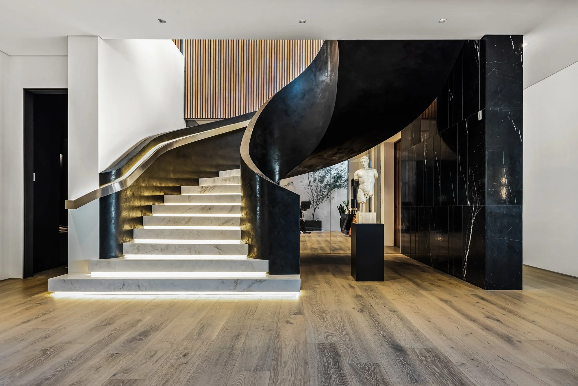 Spiral stair with glowing marble treads, dark metal rail, and black marble wall under cool light.