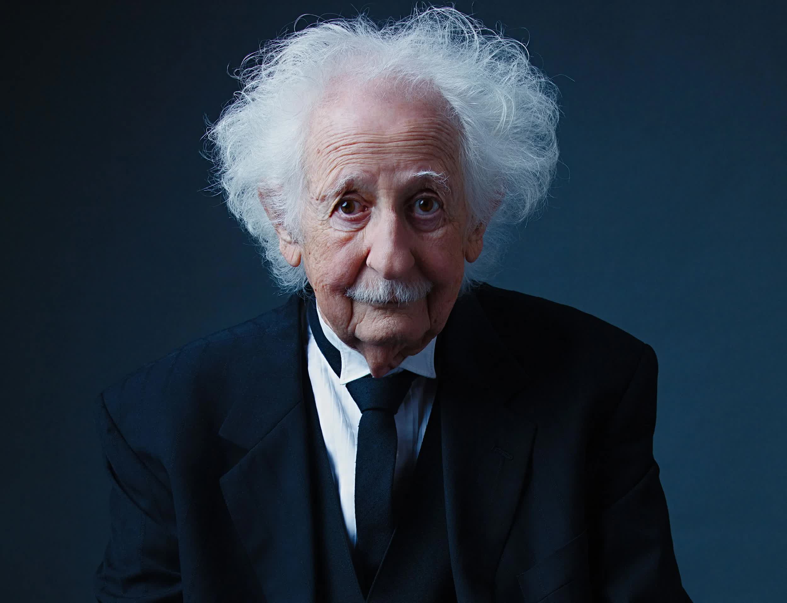 Close studio portrait of an older man with wild white hair in a black suit against a dark backdrop.