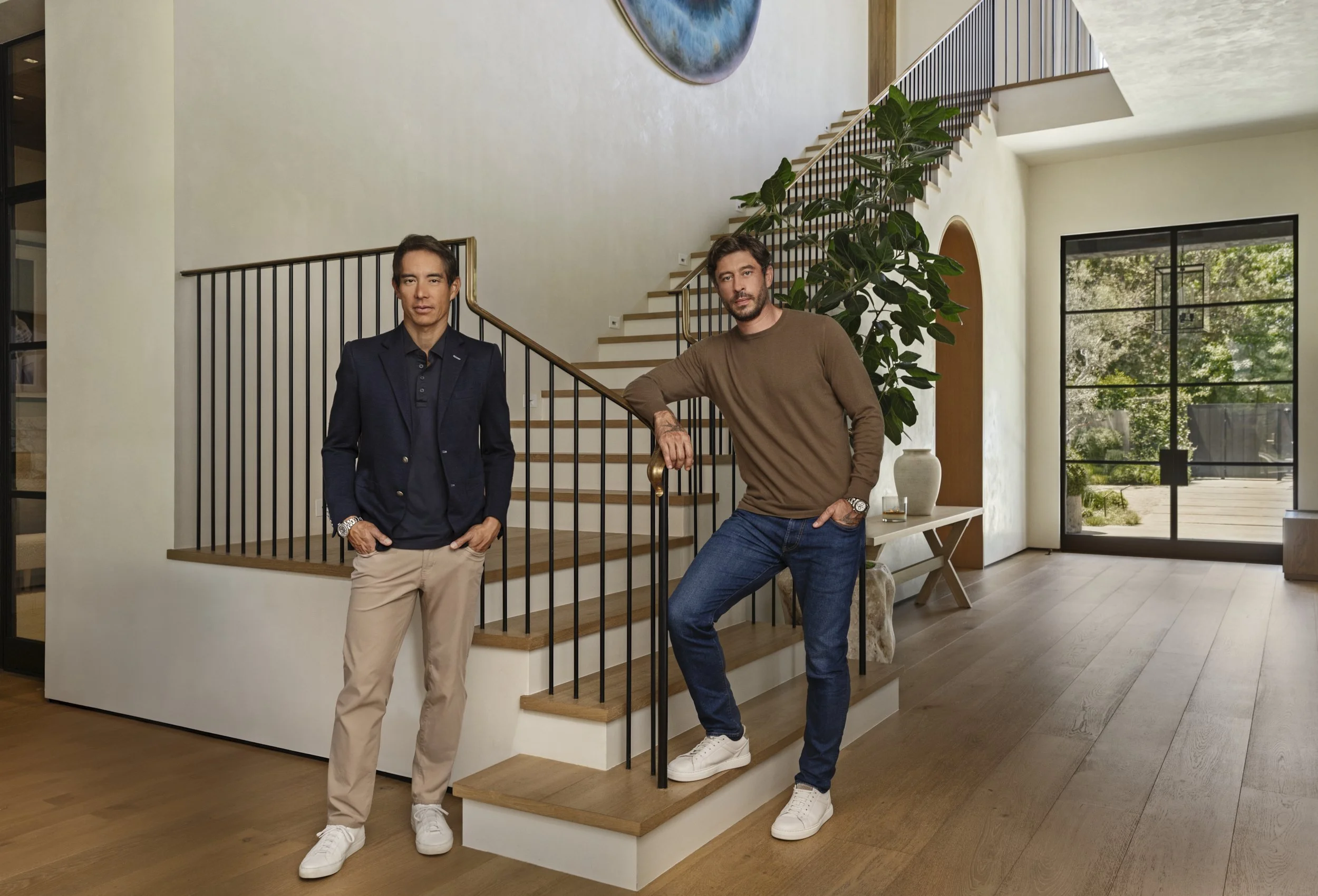 Portrait of Blair Chang and Sam Palmer by a wood stair in a sunlit modern foyer.