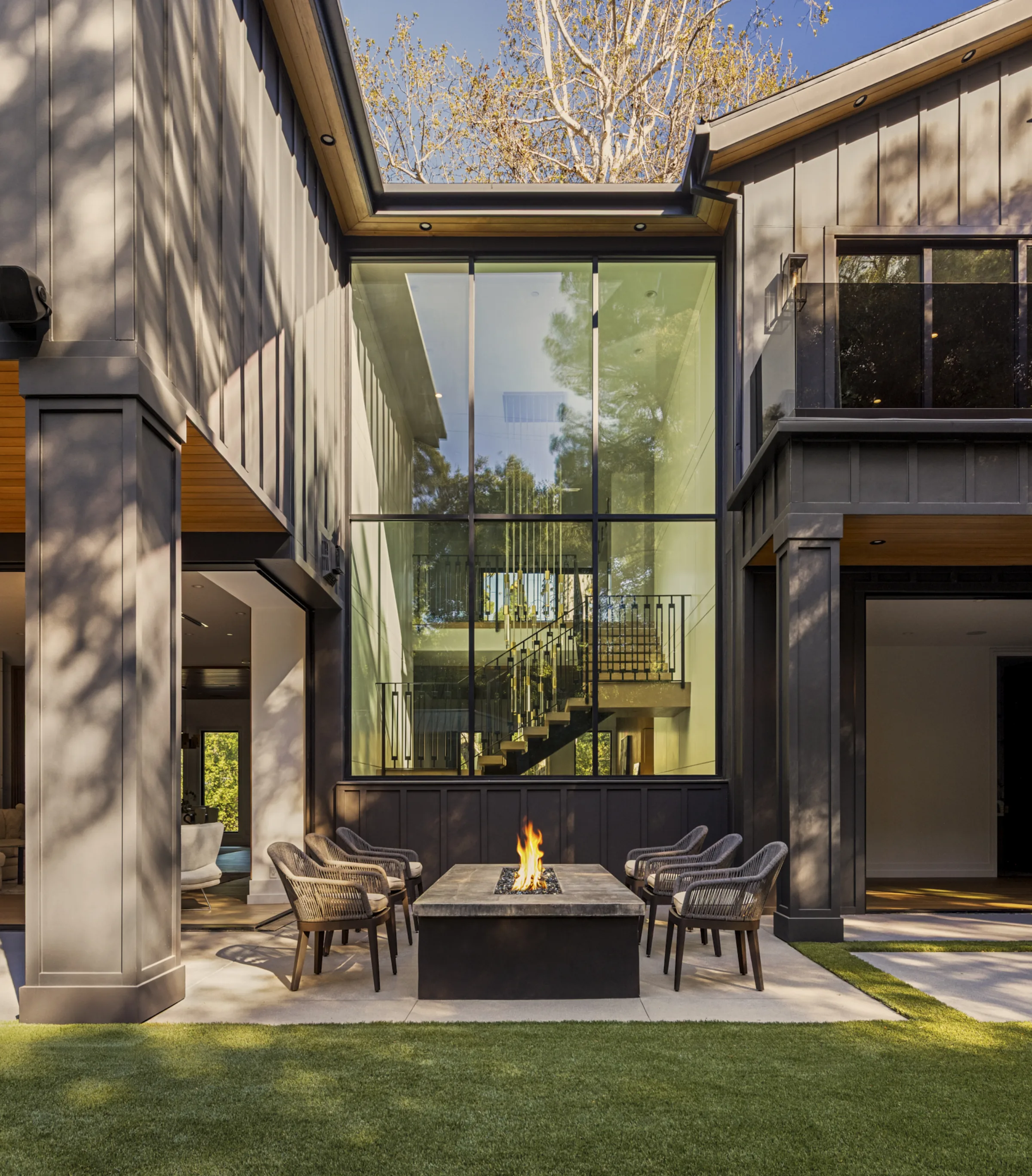 Courtyard fire pit with woven chairs before a two-story glass stair wall and dark farmhouse volumes.